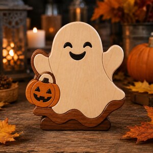 Puede incluir: Una figura de fantasma de madera con un cuerpo de color claro y una cara sonriente, que sostiene una pequeña calabaza naranja tallada. El fantasma se apoya sobre una base de madera marrón oscuro, creando una pieza decorativa en capas.