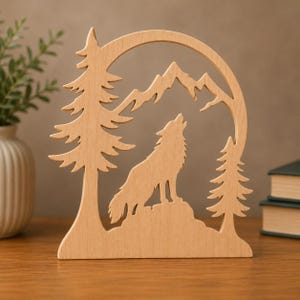 May include: A light-colored wooden cutout featuring a silhouette of a wolf howling on a rock, framed by two pine trees and a mountain range. The design is carved from a single piece of wood with a smooth finish and a natural light brown grain.