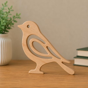 Könnte beinhalten: Eine hellbraune Vogelfigur aus Holz mit einem ausgeschnittenen Design, das die Form des Flügels und des Körpers nachzeichnet. Der Vogel steht im Profil mit einem kreisförmigen Augenloch und einer glatten Oberfläche aus natürlichem Holz.