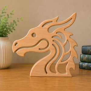 May include: A light brown wooden cutout in the shape of a dragon head, featuring a smooth finish and intricate, flowing lines that define the snout, eye, and neck crest. The object is a flat, stylized representation of a mythical creature.