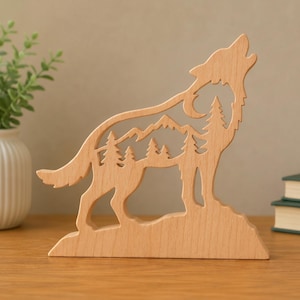 May include: A light-colored wooden cutout sculpture shaped like a howling wolf. The interior of the wolf silhouette features a detailed laser-cut scene depicting a mountain range and a forest of pine trees, all set on a flat wooden base.