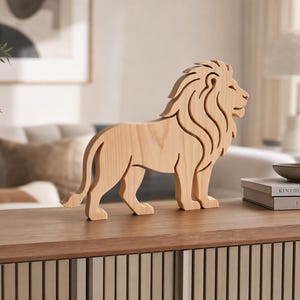 Puede incluir: Una figura de león de madera de color claro en vista de perfil. La escultura presenta líneas suaves y limpias que definen la melena, el cuerpo y la cola del león, mostrando la veta natural de la madera. La figura se muestra de pie.