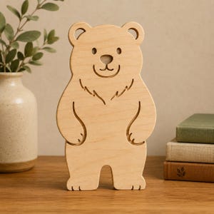 Könnte beinhalten: Eine hellfarbene Bärenfigur aus Holz mit glatter Oberfläche. Der Bär ist stehend dargestellt und besitzt abgerundete Ohren, ein einfach geschnitztes Gesicht mit Augen, Nase und lächelndem Mund sowie eingravierte Linien für die Fellstruktur.