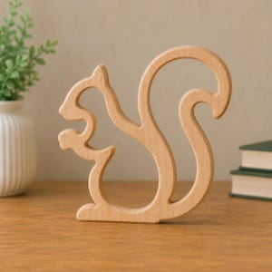 May include: A light-colored wooden cutout in the shape of a squirrel. The figure is depicted in profile, showing the outline of the animal with a curved tail and a small object held in its front paws. The wood has a smooth, natural grain finish.