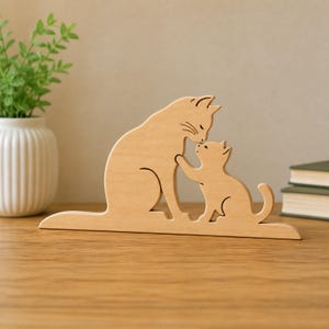 Op de afbeelding: Een lichtgekleurd houten uitgesneden sculptuur met een grote kat en een klein kitten die tegenover elkaar staan. De volwassen kat bevindt zich aan de linkerkant, terwijl het kitten aan de rechterkant staat, met hun hoofden tegen elkaar aan.