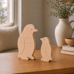 May include: Two light-coloured wooden penguin figurines standing side by side. The larger penguin is on the left and the smaller penguin is on the right. Both figures feature smooth, rounded edges and a simple carved line indicating a wing.