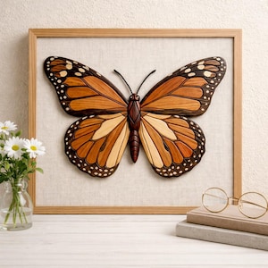 May include: A framed wooden butterfly art piece. The butterfly features intricate details with brown, orange, and cream-colored wood pieces. The artwork is set against a neutral fabric background within a light brown wooden frame.