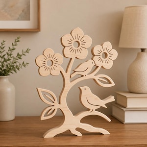 Könnte beinhalten: Eine helle Holzschnitzerei in Form eines stilisierten Baumes mit drei fünfblättrigen Blüten und zwei Vögeln, die auf den Ästen sitzen. Das Design umfasst ausgeschnittene Blütenmitten sowie lineare Gravuren auf den Blütenblättern und Vögeln.