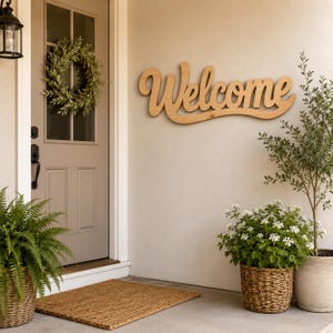 May include: A light brown wooden wall sign featuring the word Welcome in a cursive script font. The sign is mounted on a beige wall next to a door with a green wreath, a brown doormat, and two potted plants in woven baskets on the porch.