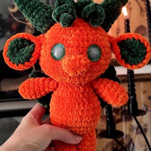 Può includere: Una creatura all'uncinetto arancione fatta a mano con accenti verdi, grandi occhi verdi e corna verdi. Il peluche ha un design stravagante ed è tenuto da una persona. Il testo "Whimsy Darkly Créations" è visibile in basso.