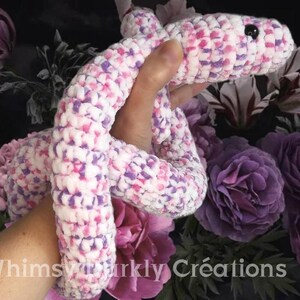 Peut inclure: Un serpent en crochet fait à la main, blanc, rose et violet. Le jouet en peluche a un œil en bouton noir et est présenté sur un fond floral. Le serpent est fait de fil doux et texturé.
