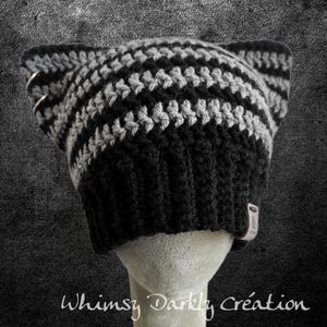 May include: A black and grey striped crochet hat with cat ears and silver hoop accents. The hat is displayed on a mannequin head. The text "Whimsy Darkly Création" is at the bottom.