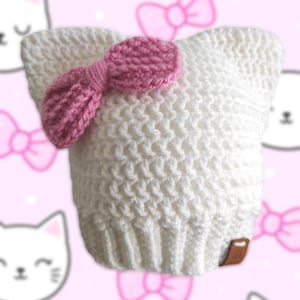 Op de afbeelding: Een witte gehaakte muts met kattenoordetails en een roze strik. De muts heeft een gestructureerd steekpatroon en een klein bruin label. De tekst "Whimsy darkly créations" is zichtbaar.