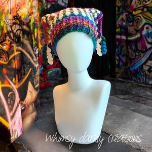 Peut inclure: Un chapeau crocheté fait main aux couleurs vives, notamment violet, bleu, vert et blanc. Le chapeau a un sommet pointu et des pompons décoratifs. Le texte "Whimsy darly creations" est visible en bas. Un article unique et artisanal.