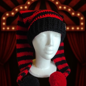 Pode incluir: Um chapéu de crochê feito à mão, com listras pretas e vermelhas, com um design longo e pontudo. O chapéu apresenta um pompom preto em uma extremidade e um pompom vermelho na outra. O texto "Whimsy darkly créations" é visível na parte inferior.