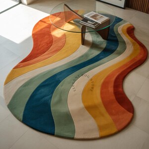 Könnte beinhalten: Ein farbenfroher, wellenförmiger Teppich mit Regenbogenmuster. Der Teppich hat Streifen in Orange, Gelb, Grün, Blau und Creme. Die Worte "ELEGANT RUG STORE" sind auf dem Teppich aufgedruckt. Ein Couchtisch aus Glas steht darauf.