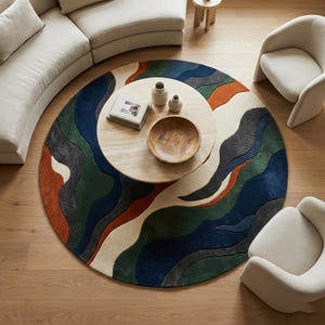 May include: A round area rug with a flowing, abstract design in shades of blue, green, orange, gray, and cream. The rug is centered in a room with a light-colored sofa, two chairs, and a coffee table.