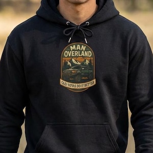 May include: Black hooded sweatshirt with a front pocket and drawstring. The sweatshirt features a graphic with the text "MAN OVERLAND" above an image of a vehicle in a mountain landscape. The text "OLD TOYOTAS DO IT BETTER" is below the image.