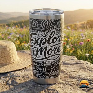 May include: Stainless steel travel tumbler with a black and white design and the words "Explore More". The tumbler has a silver lid and base. The background shows a straw hat and a field of wildflowers.