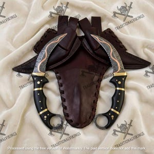 May include: Two curved karambit knives with ornate blades and black handles with gold accents. The knives are displayed with a brown leather sheath. The blades have a copper-coloured design. The handles have a finger ring.