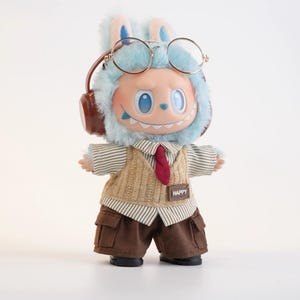 Puede incluir: Una mu&ntilde;eca caprichosa con una capucha azul con orejas de conejo, gafas redondas y auriculares. Viste una camisa a rayas, un chaleco de punto beige, una corbata roja y pantalones cargo marrones. La mu&ntilde;eca tiene una sonrisa amplia y un parche "HAPPY".