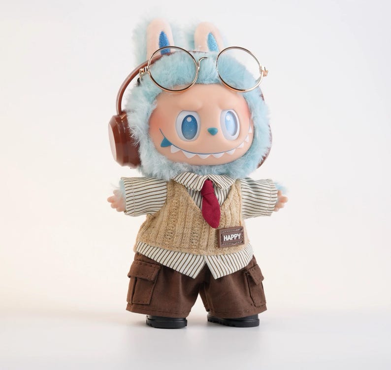 Puede incluir: Una mu&ntilde;eca caprichosa con una capucha azul con orejas de conejo, gafas redondas y auriculares. Viste una camisa a rayas, un chaleco de punto beige, una corbata roja y pantalones cargo marrones. La mu&ntilde;eca tiene una sonrisa amplia y un parche "HAPPY".