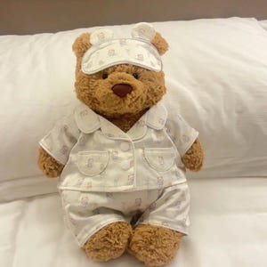 May include: A brown teddy bear wearing a white pajama set with a light blue pattern and a matching sleep mask. The bear is sitting on a white surface. The pajamas include a button-up shirt and shorts.