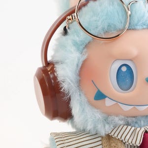Puede incluir: Primer plano de una figura caprichosa con una cabeza peluda azul claro, que lleva gafas con montura dorada y auriculares marrones. La figura tiene una cara rosa claro, ojos azules y un atuendo cosido. El tema general es un personaje lindo y de dibujos animados.