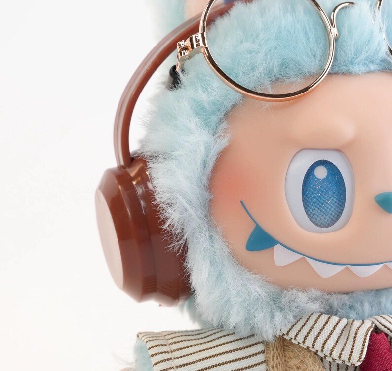 Puede incluir: Primer plano de una figura caprichosa con una cabeza peluda azul claro, que lleva gafas con montura dorada y auriculares marrones. La figura tiene una cara rosa claro, ojos azules y un atuendo cosido. El tema general es un personaje lindo y de dibujos animados.