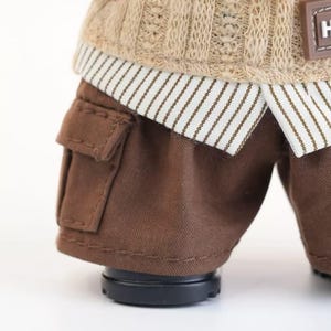 Puede incluir: Primer plano de un atuendo de mu&ntilde;eca con pantalones cortos cargo marrones con un bolsillo, un su&eacute;ter de punto beige y una camisa con cuello con un patr&oacute;n de rayas. El atuendo se completa con zapatos negros.