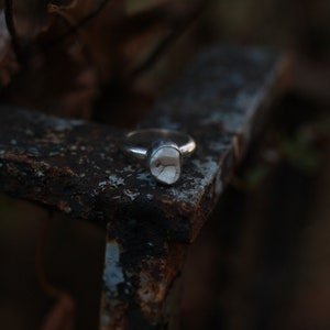 Op de afbeelding: Een zilveren ring met een unieke, onregelmatig gevormde, gebroken witte steen. De ring ligt op een verweerd, roestig metalen oppervlak, met een vage natuurlijke achtergrond.