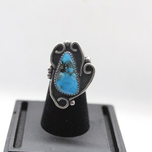 Pode incluir: Um anel de prata com uma grande pedra turquesa oval com matriz escura. A pedra é engastada em uma moldura de prata ornamentada com detalhes em espiral. O anel é exibido em um suporte preto sobre um fundo branco.