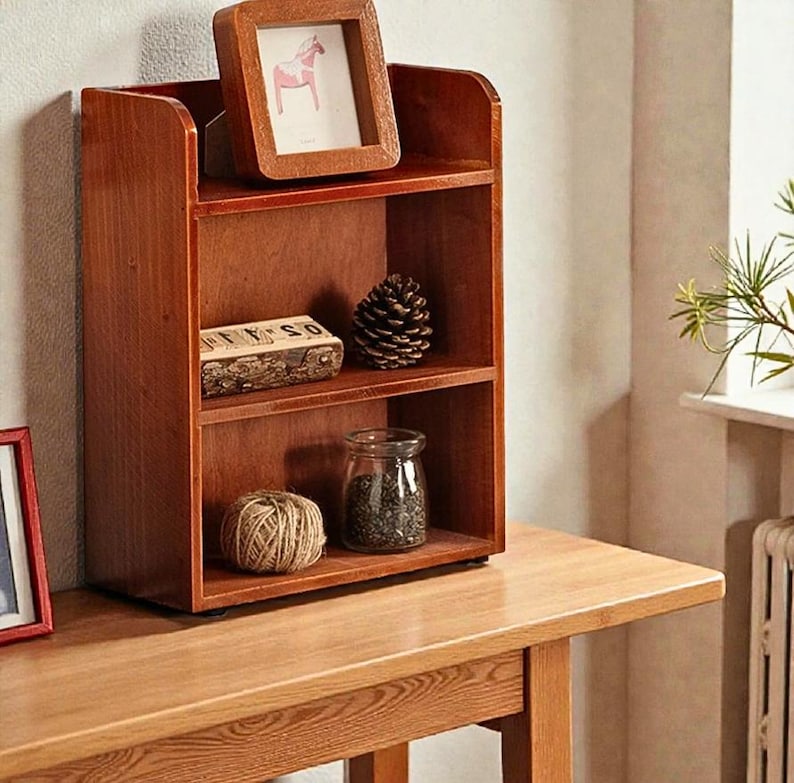May include: A three-tiered wooden shelf unit with a warm brown finish. The top shelf holds a framed picture of a horse. Other shelves display a pine cone, a small jar, and a ball of twine. The unit sits on a wooden table.