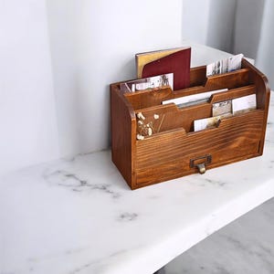 May include: A wooden desk organizer with multiple compartments and a drawer. The organizer is stained a warm brown color and contains various cards and a small book. A small metal pull is on the drawer.
