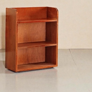 May include: A small, two-shelf wooden bookcase with a warm, reddish-brown finish. The bookcase has a simple, rectangular design with rounded edges on the top shelf. The shelves are open, providing storage space. The bookcase is suitable for books or decorative items.