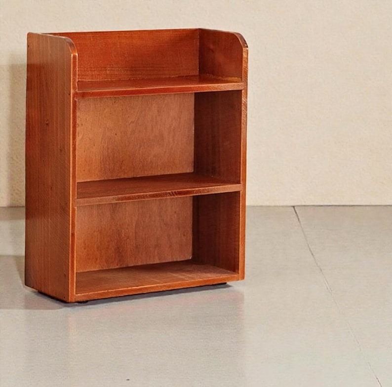 May include: A small, two-shelf wooden bookcase with a warm, reddish-brown finish. The bookcase has a simple, rectangular design with rounded edges on the top shelf. The shelves are open, providing storage space. The bookcase is suitable for books or decorative items.