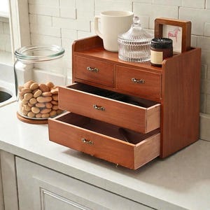 May include: A wooden storage chest with three drawers, two small drawers on top, and a shelf. The chest is made of brown wood and has decorative metal drawer pulls. A glass jar with nuts, a white mug, and a candle sit nearby.