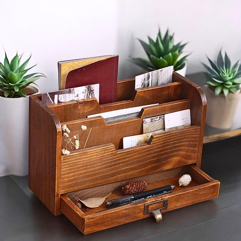 May include: A wooden desk organizer with multiple compartments and a drawer. The organizer is stained a warm brown color and contains various items, including cards, a book, and pens. Decorative plants are in the background.