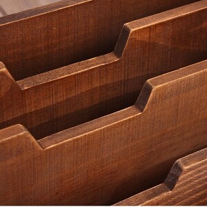 May include: Close-up of a wooden organizer with multiple compartments. The wood has a warm, brown tone and visible grain. Each compartment has a stepped design, creating a tiered effect. The organizer appears to be handcrafted.