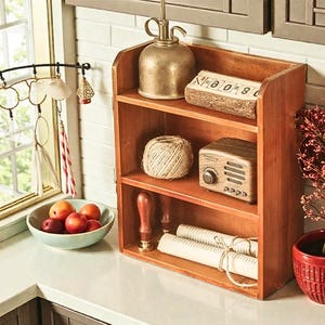 May include: A wooden shelf unit with three levels displays vintage decor. Items include a metal watering can, a wooden block with numbers, a ball of twine, a miniature radio, and rolled paper. A bowl of fruit and hanging decorations are also visible.