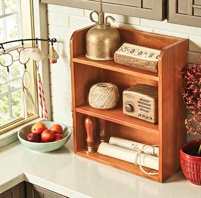 May include: A wooden shelf unit with three levels displays vintage decor. Items include a metal watering can, a wooden block with numbers, a ball of twine, a miniature radio, and rolled paper. A bowl of fruit and hanging decorations are also visible.