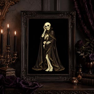 May include: A framed wall art print featuring a cream-coloured skeleton wearing a long, flowing black cape with intricate lace trim. The skeleton is depicted in a standing pose with one hand resting near its face, set against a solid black background.