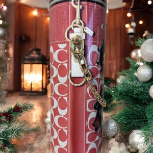 May include: A red and white patterned tumbler with a gold-coloured keychain attached. The tumbler has a leather-like texture and a silver rim. The design includes the letter "C" repeated in a pattern.