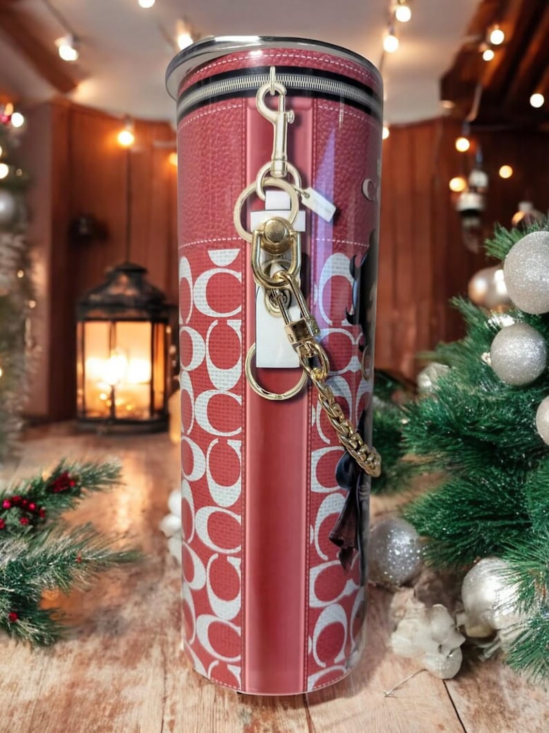 May include: A red and white patterned tumbler with a gold-coloured keychain attached. The tumbler has a leather-like texture and a silver rim. The design includes the letter "C" repeated in a pattern.