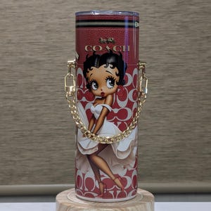 May include: A red and white Coach tumbler with a Betty Boop graphic. The tumbler has a gold chain handle and a red leather-like texture. The Coach logo is visible. The tumbler is cylindrical and sits on a wooden base.