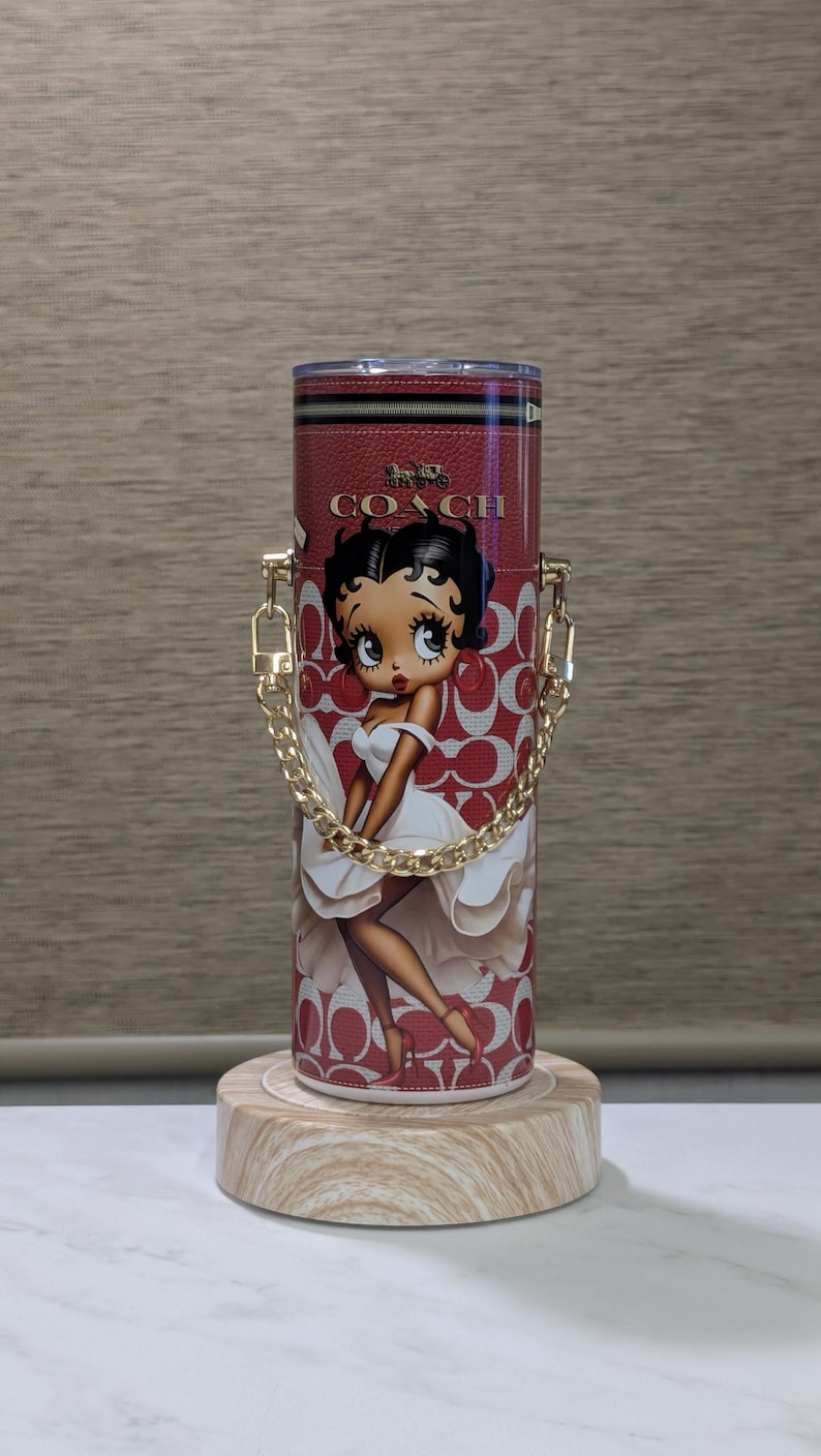 May include: A red and white Coach tumbler with a Betty Boop graphic. The tumbler has a gold chain handle and a red leather-like texture. The Coach logo is visible. The tumbler is cylindrical and sits on a wooden base.