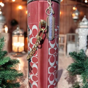 May include: A red and white Coach tumbler with a zipper design at the top. The tumbler features a repeating pattern of the Coach logo and includes a gold chain and a Coach tag. The background is blurred with Christmas decorations.
