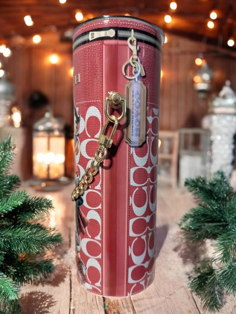 May include: A red and white Coach tumbler with a zipper design at the top. The tumbler features a repeating pattern of the Coach logo and includes a gold chain and a Coach tag. The background is blurred with Christmas decorations.