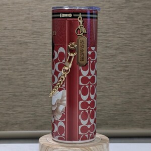 May include: A red and white Coach tumbler with a floral design and the Coach logo. The tumbler has a gold chain and a zipper design. The tumbler is cylindrical and sits on a wooden stand.