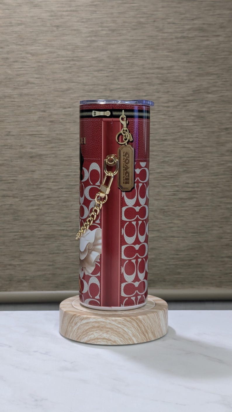 May include: A red and white Coach tumbler with a floral design and the Coach logo. The tumbler has a gold chain and a zipper design. The tumbler is cylindrical and sits on a wooden stand.